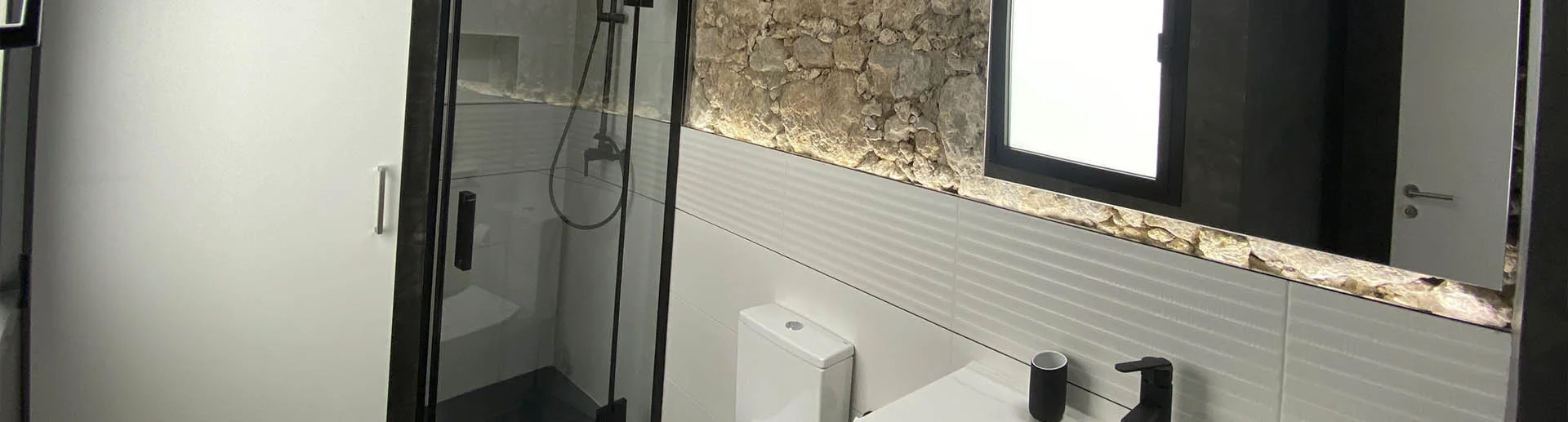 Baño reformado con pared de piedra natural, ducha con mampara de vidrio, sanitarios blancos y grifería negra. Proyecto moderno y funcional. Foto propiedad de Vigasa.
