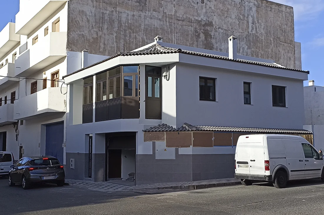 Casa de esquina reformada con fachada blanca y zócalo gris, techo de tejas y cerramiento acristalado en planta alta. Proyecto de construcciones y reformas integrales. Foto propiedad de Vigasa.
