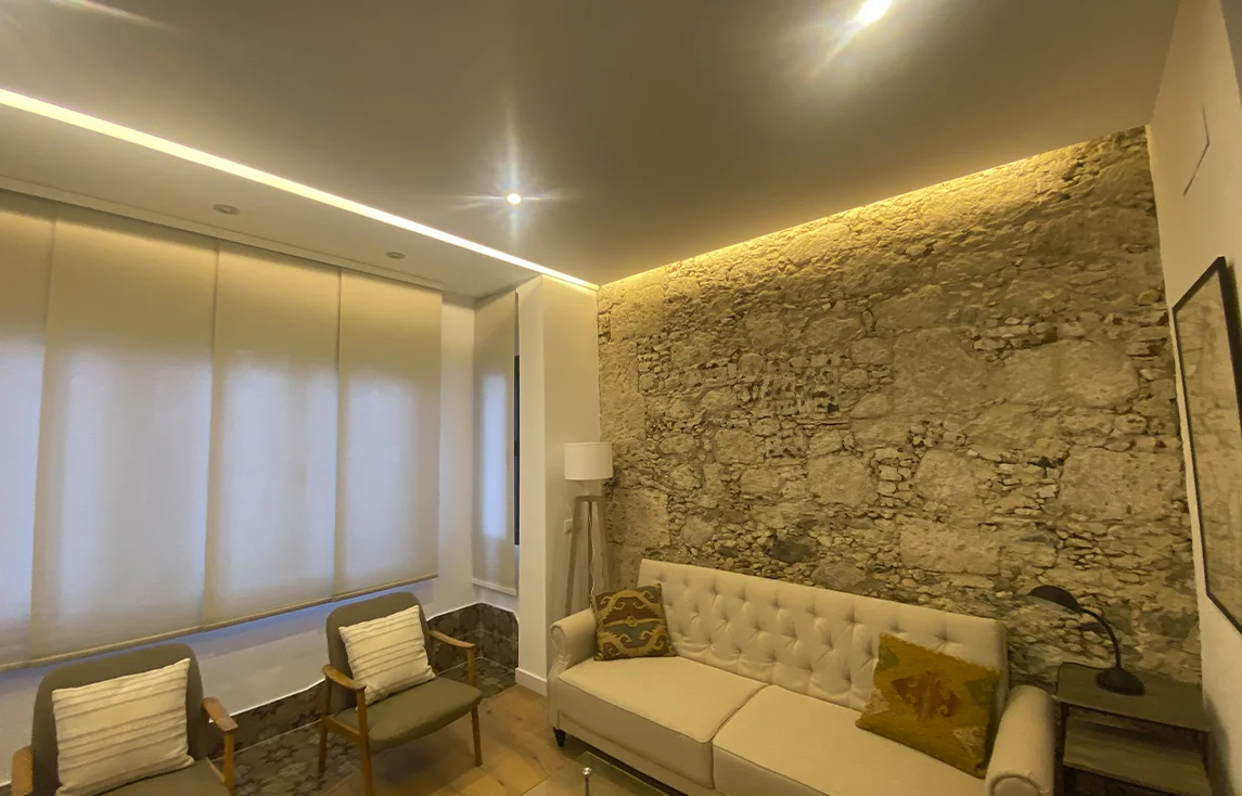 Sala de estar reformada con diseño moderno y detalles rústicos, destacando una pared de piedra vista restaurada con iluminación LED indirecta. La estancia incluye un sofá beige capitoné, dos sillones individuales con cojines claros, lámpara de pie, cortinas enrollables translúcidas y techo con iluminación empotrada. Proyecto realizado por Vigasa Proyectos y Obras, especializado en construcciones y reformas y servicios de albañilería.