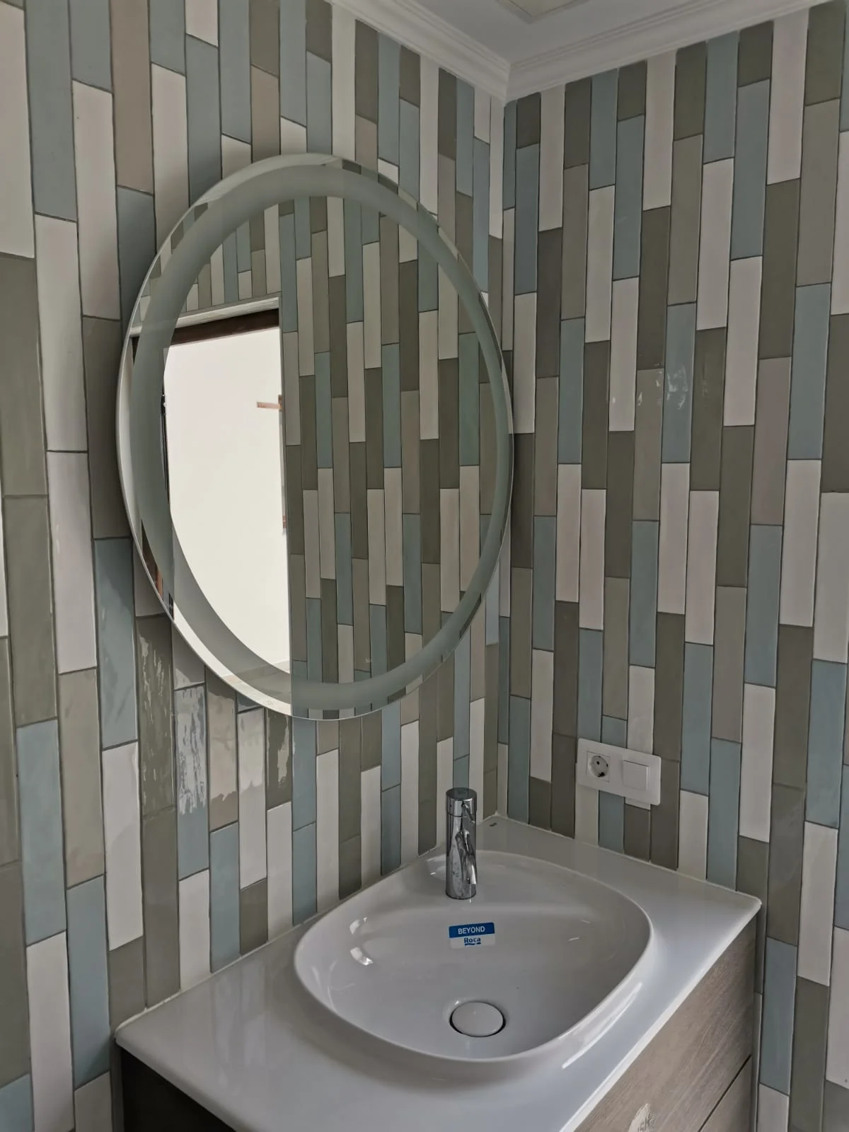 Lavabo moderno con espejo redondo en baño con azulejos verticales de colores verdes, blancos y grises. Foto propiedad de Vigasa.