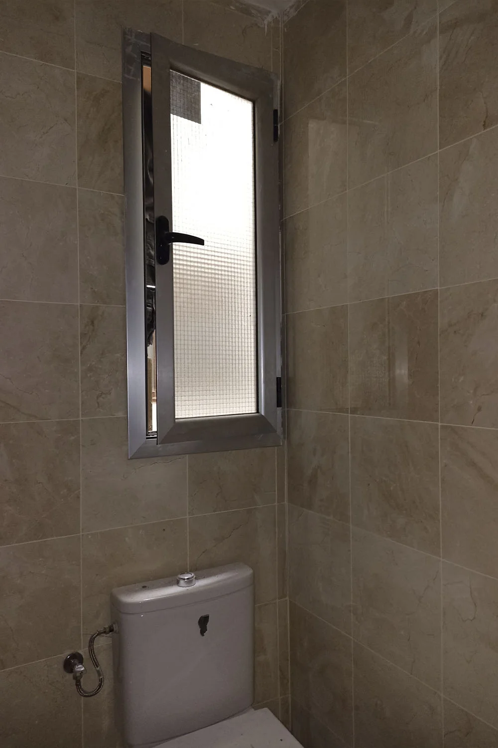 Baño en proceso de reforma con inodoro instalado bajo una ventana abatible de aluminio y paredes revestidas en mármol beige. Foto propiedad de Vigasa.