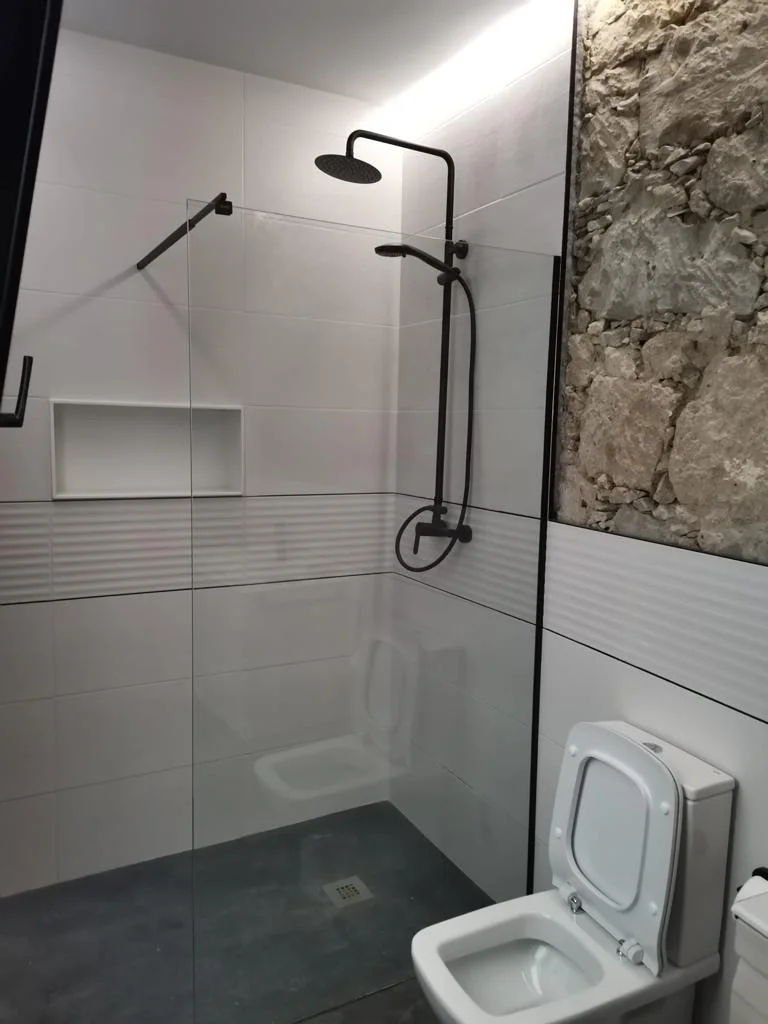 Baño reformado con ducha de obra, grifería negra, pared de piedra natural y revestimientos cerámicos blancos. Foto propiedad de Vigasa.