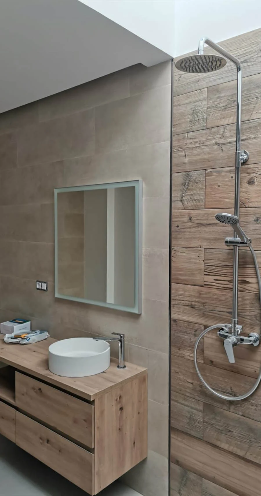 Baño en proceso de reforma con mueble de madera, lavabo sobre encimera, ducha con efecto lluvia y revestimientos cerámicos tipo madera. Foto propiedad de Vigasa