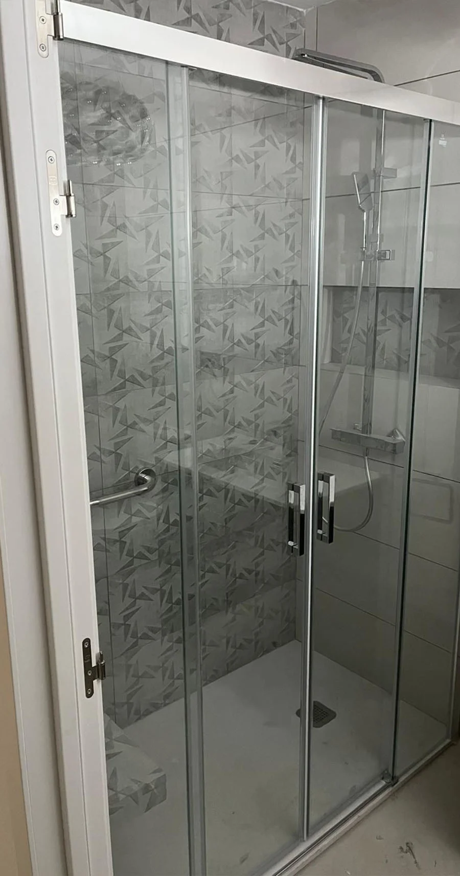 Ducha reformada con mampara de cristal corredera, banco de obra, grifería moderna y azulejos con diseño geométrico gris. Foto propiedad de Vigasa.