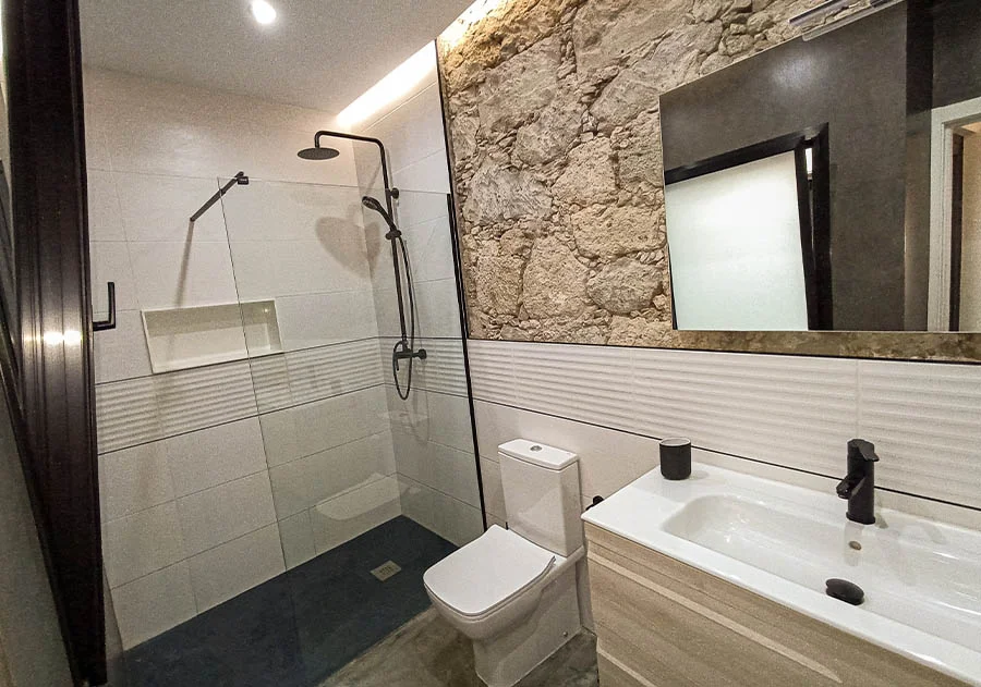 Baño reformado con ducha a ras de suelo, grifería negra, pared de piedra natural y acabados modernos en cerámica blanca. Foto propiedad de Vigasa.