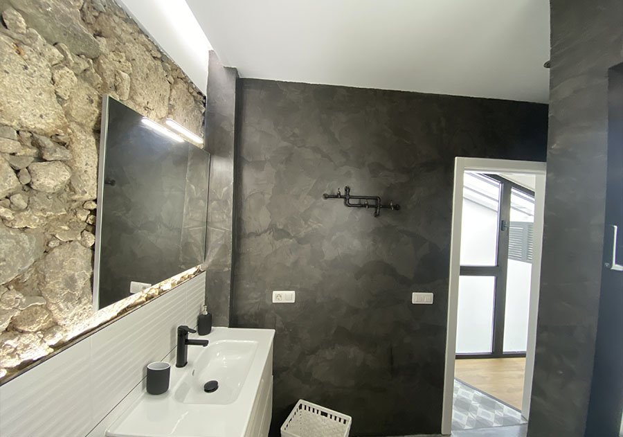 Baño reformado en Las Palmas con pared de piedra natural, lavabo blanco con grifería negra y pared oscura de acabado pulido. Foto propiedad de Vigasa.